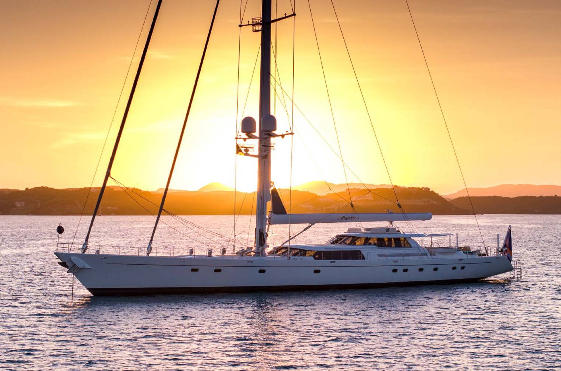 A large white sailboat rests on calm water in the foreground, silhouetted against a vibrant orange sunset over distant hills in the background.