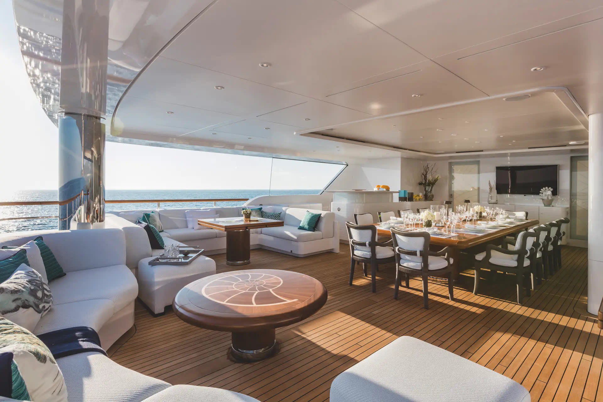 A luxurious yacht deck features a white lounge area and a round wooden table in the foreground, with a formal dining set and a view of the ocean in the background.