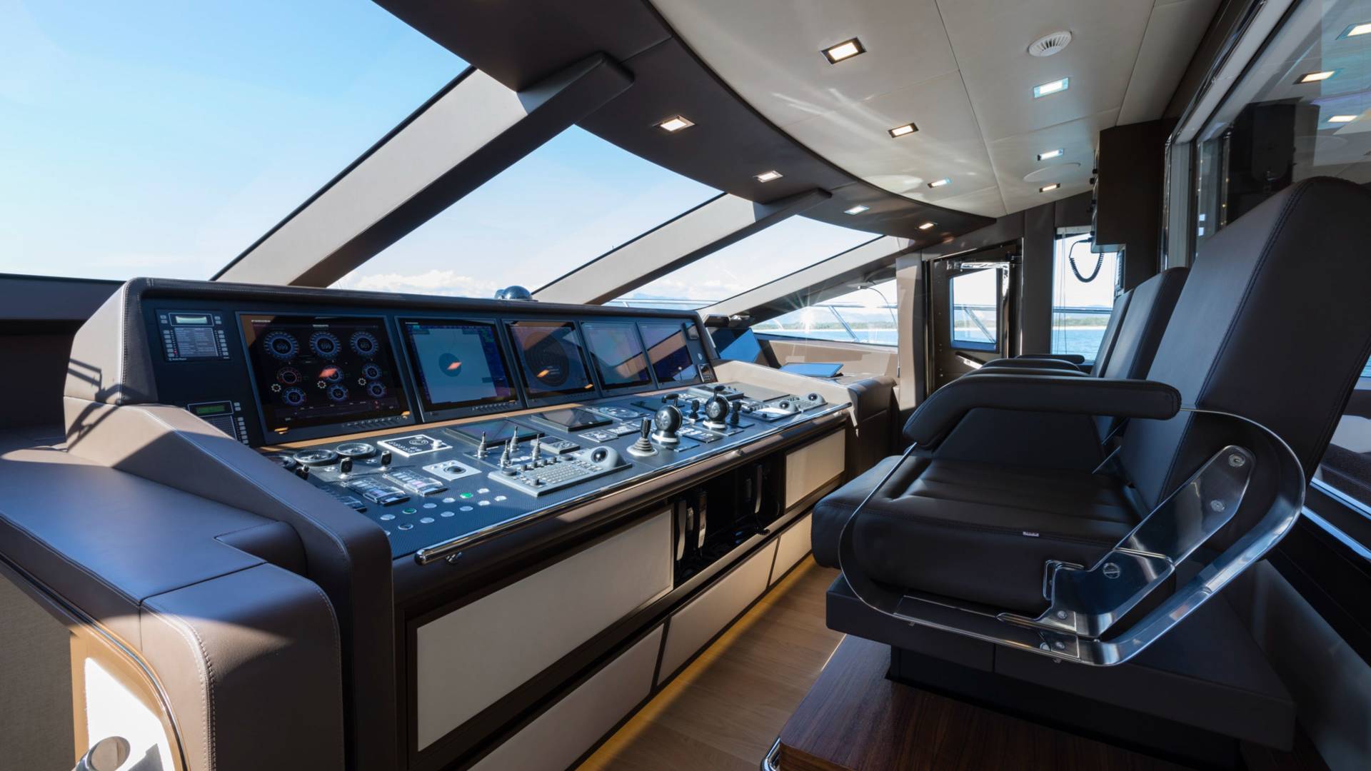 A high-tech boat cockpit features a complex control panel and leather seats in the foreground, with a bright blue sky visible through the slanted windows in the background.