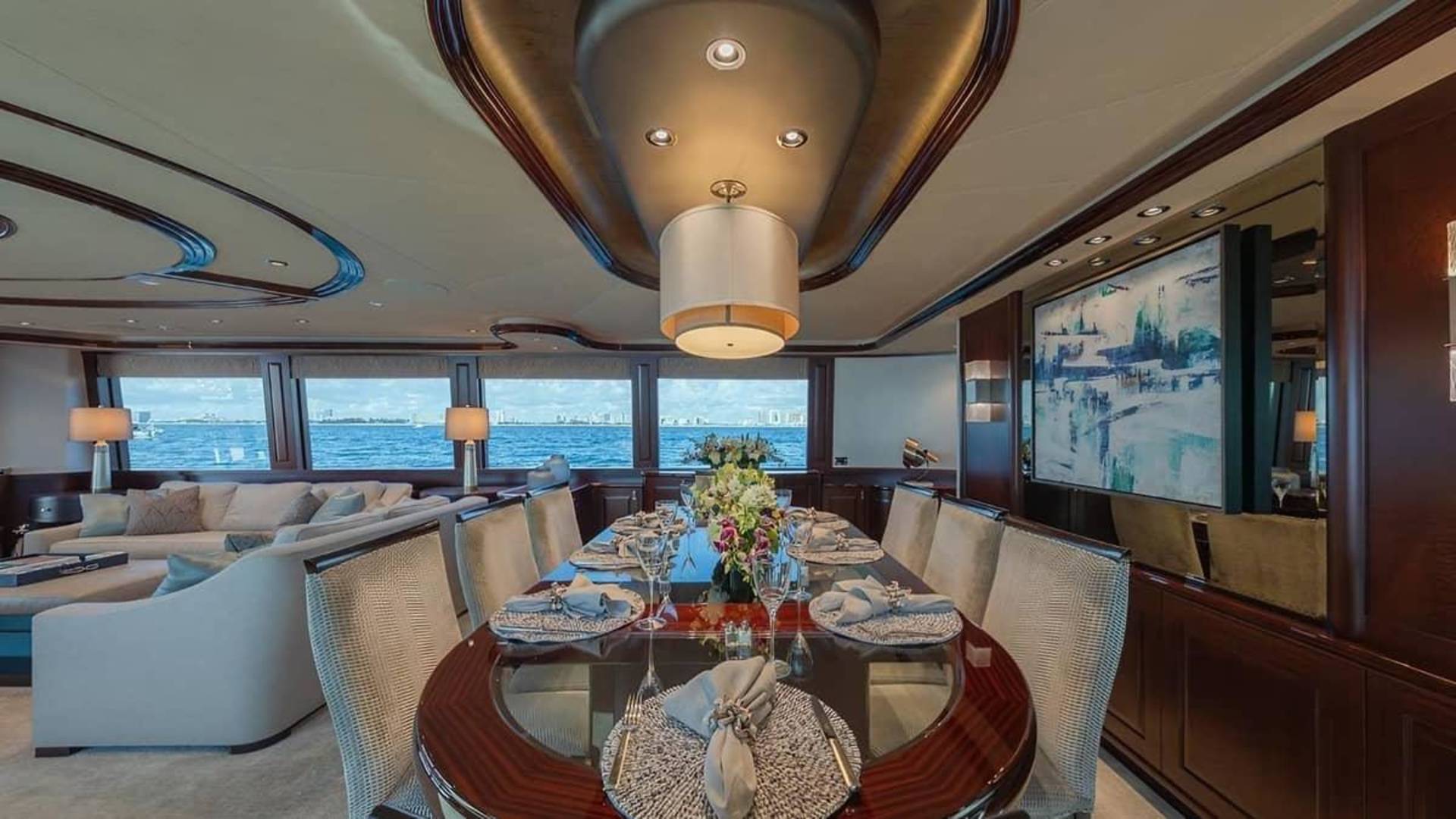 An elegant dining table set for a meal occupies the foreground of a luxury yacht interior, with a plush lounge area and ocean views visible in the background.