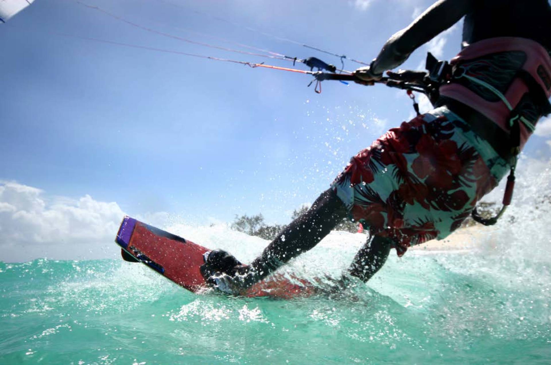 A kitesurfer in floral shorts glides across turquoise water under a bright blue sky.