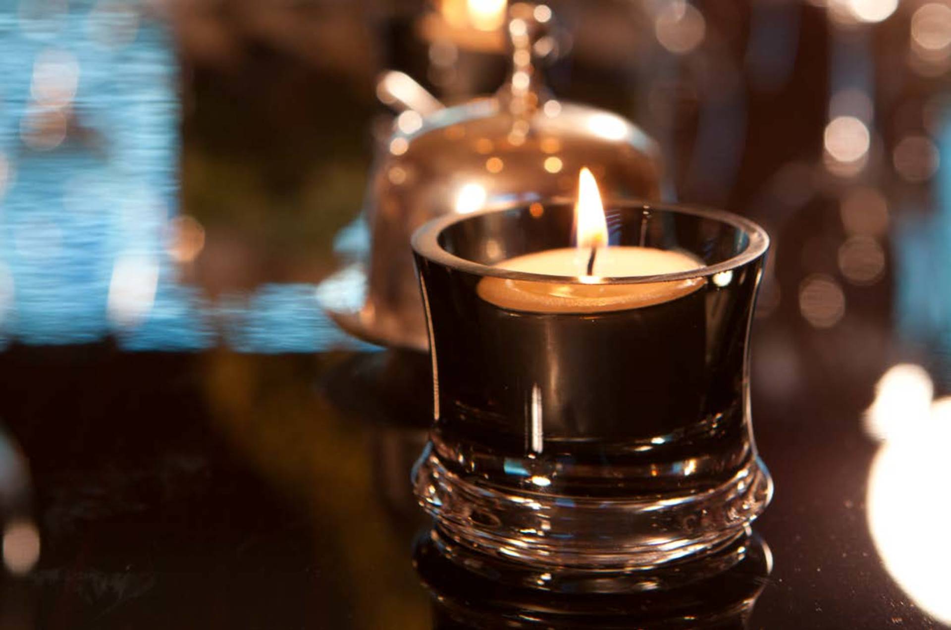A lit tea light candle in a dark glass holder sits in the foreground against a blurred background of warm lights and a silver cloche.