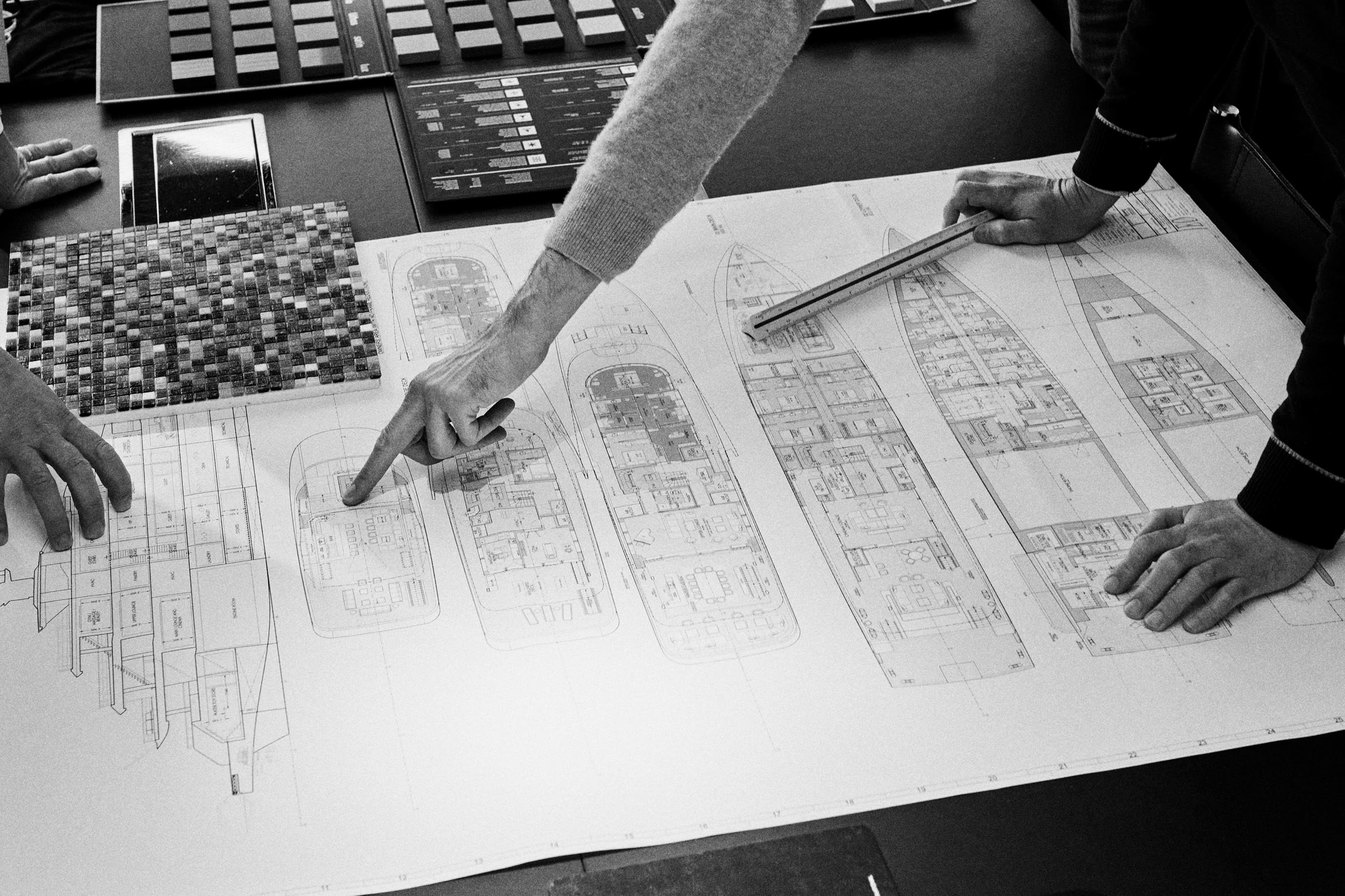The image depicts a person's hands working on architectural plans or blueprints, with a calculator and mosaic tiles visible in the background.