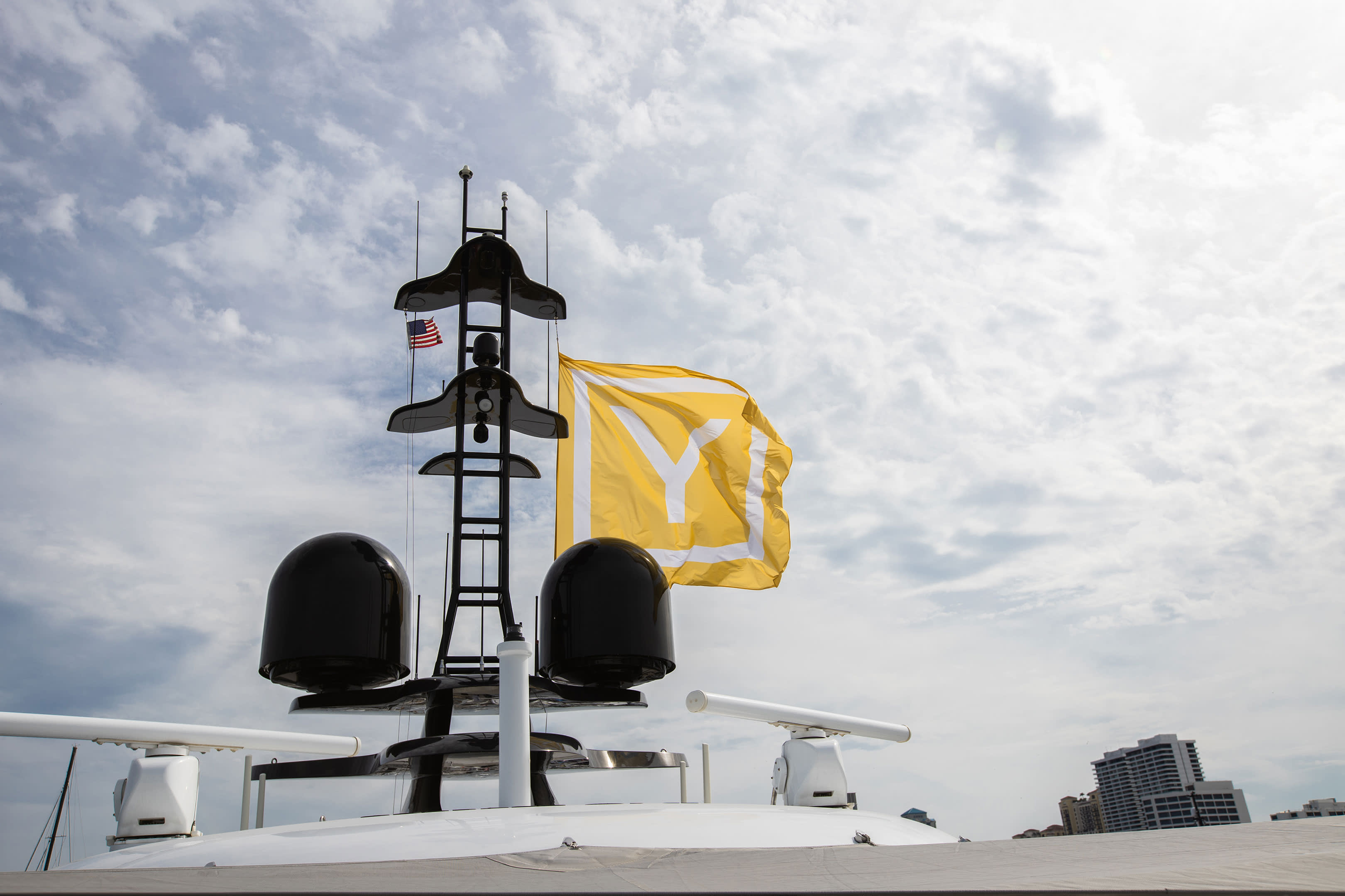 A large yellow flag with a white "Y" billows in the wind above a yacht's radar array against a cloudy sky.