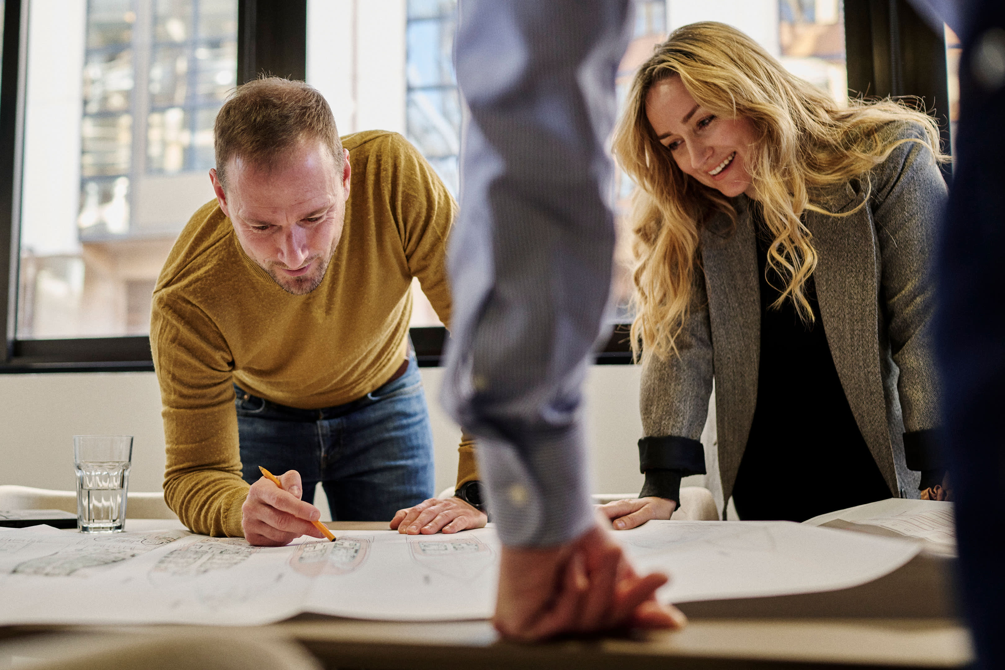 Two people, a man and a woman, are leaning over a table and examining some documents or plans together in what appears to be an office or workspace setting.