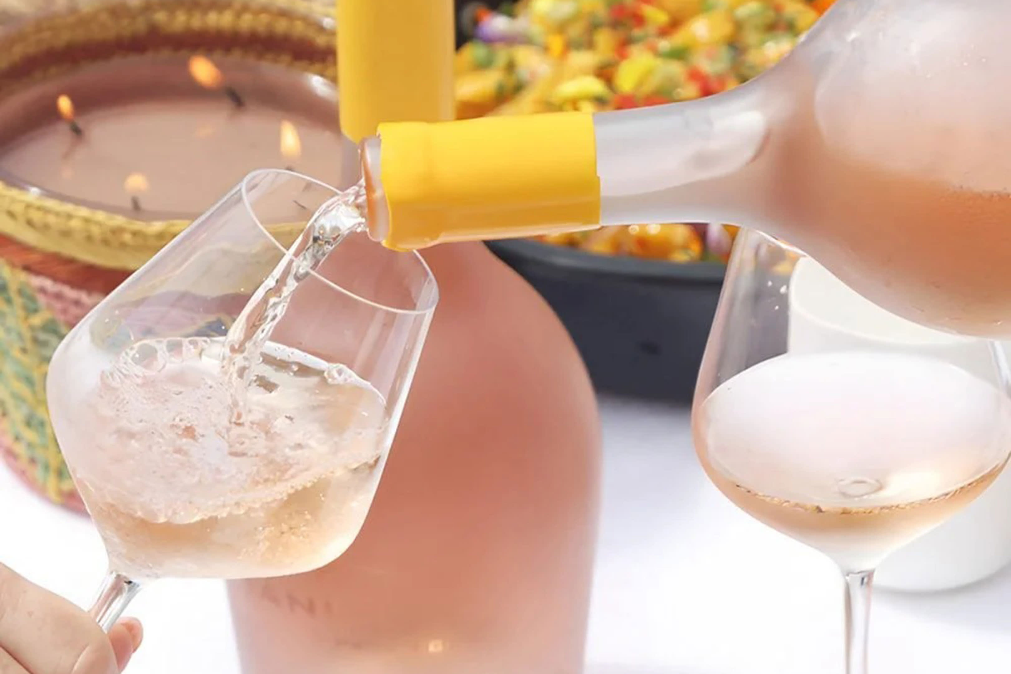 Rosé wine is poured from a bottle into a glass in the foreground, with a lit candle and a bowl of food in the blurred background.