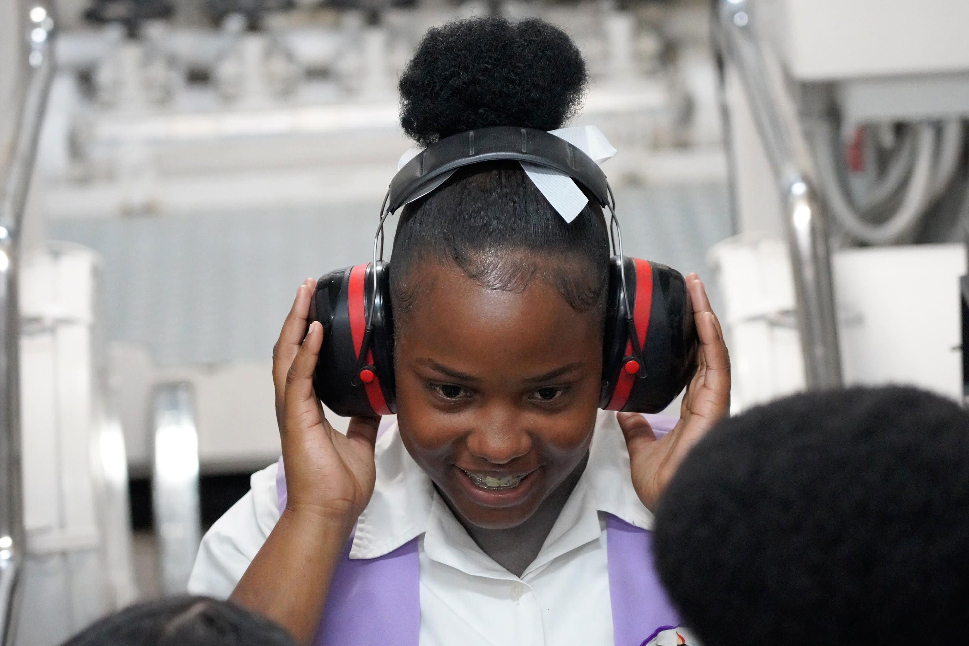 A young girl in the foreground wears large earmuffs while looking at something in the background.