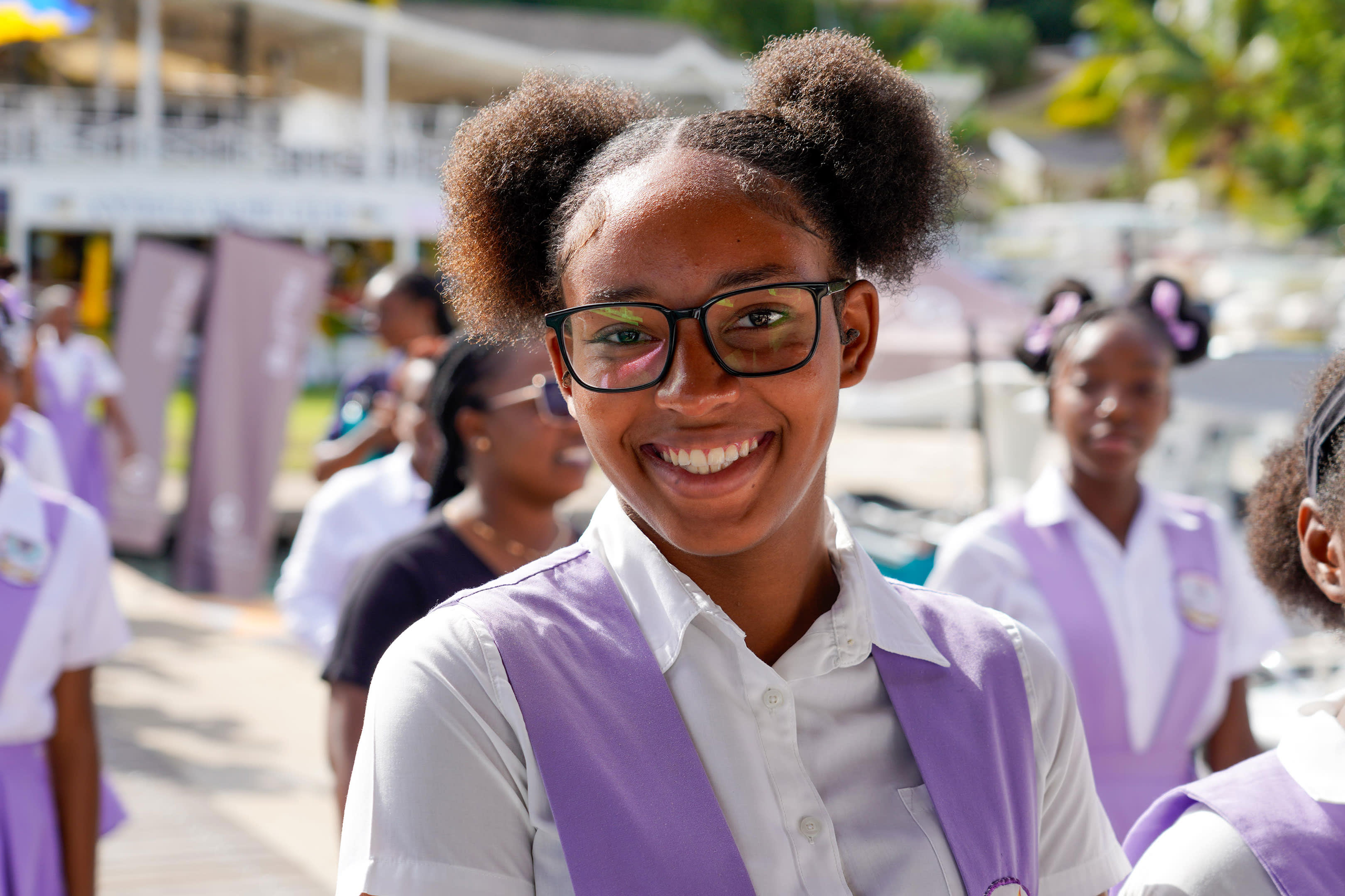 A smiling girl in glasses and a purple vest is in the foreground, with other children in similar uniforms in the blurred background.