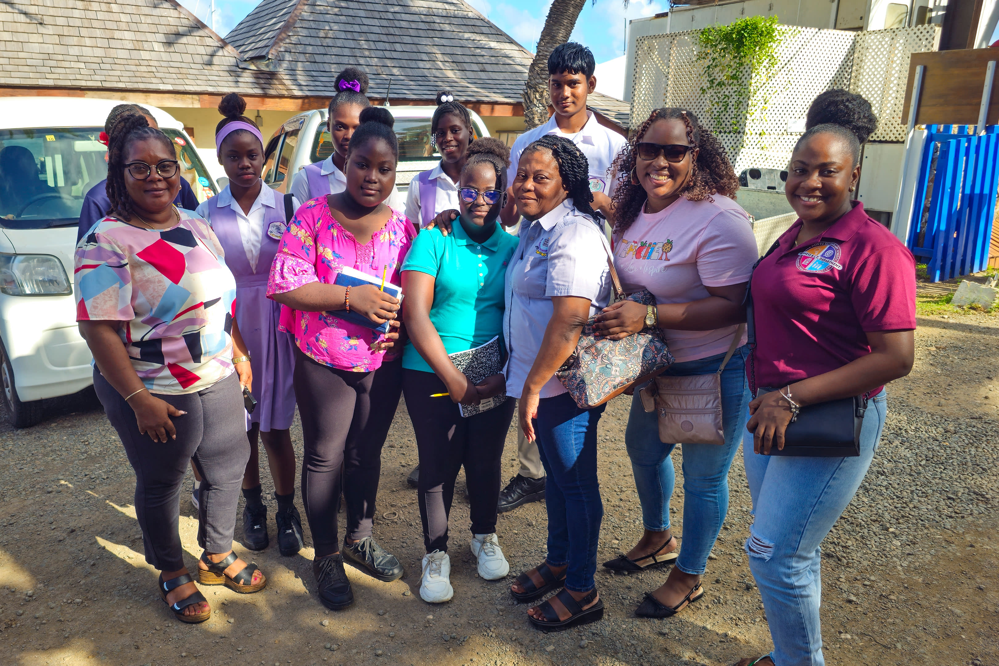 A group of smiling people, including adults and teenagers, pose for a photo in front of a white van and a building.