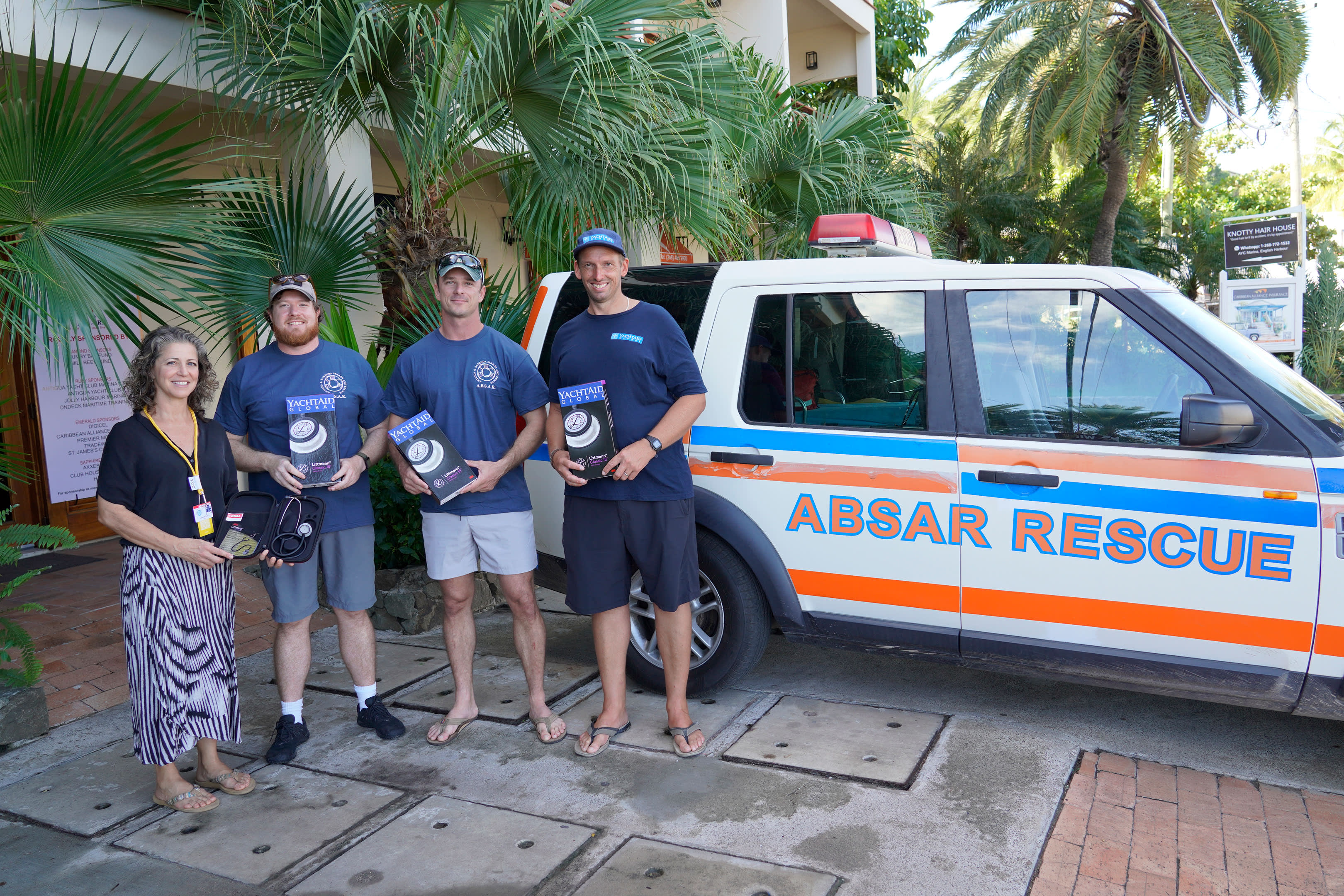 Four people stand in front of an ABSAR RESCUE vehicle with lush green foliage in the background.