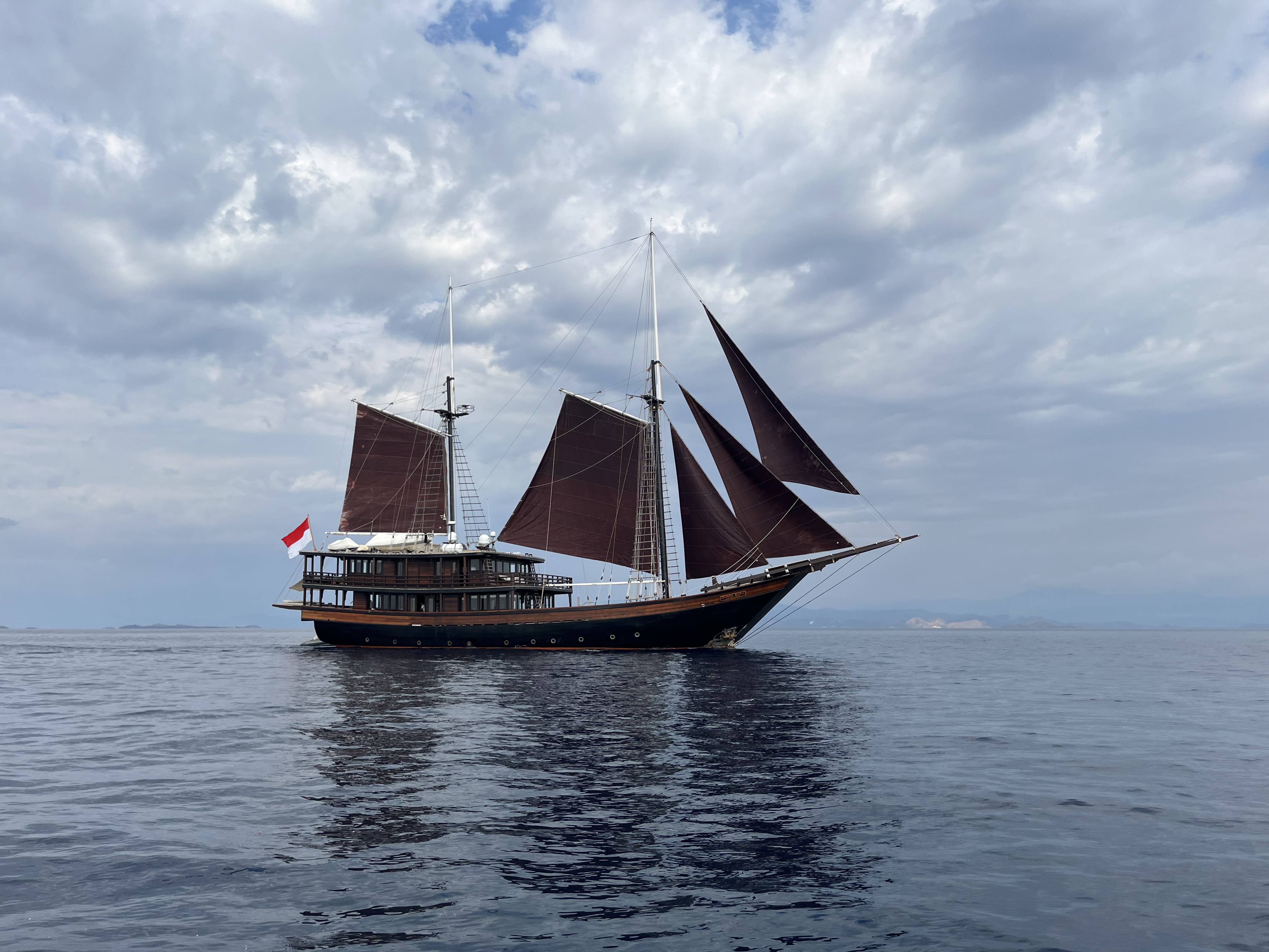 A large, traditional sailing ship with red sails and a wooden hull floats on a calm, blue ocean under a cloudy sky.