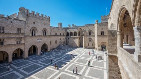 Tourists explore the vast courtyard of the Palace of the Grand Master of the Knights of Rhodes under a clear blue sky.