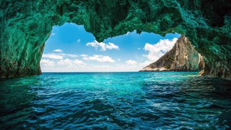 Crystal-clear turquoise waters beckon from the mouth of a sea cave, revealing a dramatic cliffside bathed in sunlight.