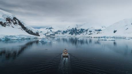 A sleek yacht navigates through a serene, icy Antarctic landscape dotted with majestic icebergs under a dramatic sky.