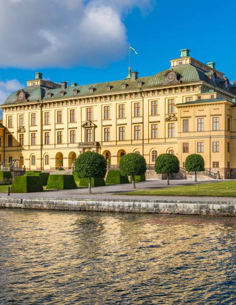 The golden facade of Drottningholm Palace reflects beautifully in the shimmering water under a bright blue sky.