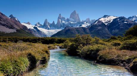 A crystal-clear river flows through a lush valley towards the majestic, snow-capped peaks of Fitz Roy.