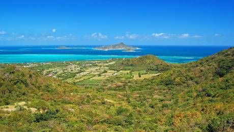 Lush green hills roll down to a turquoise sea dotted with distant islands under a brilliant blue sky.