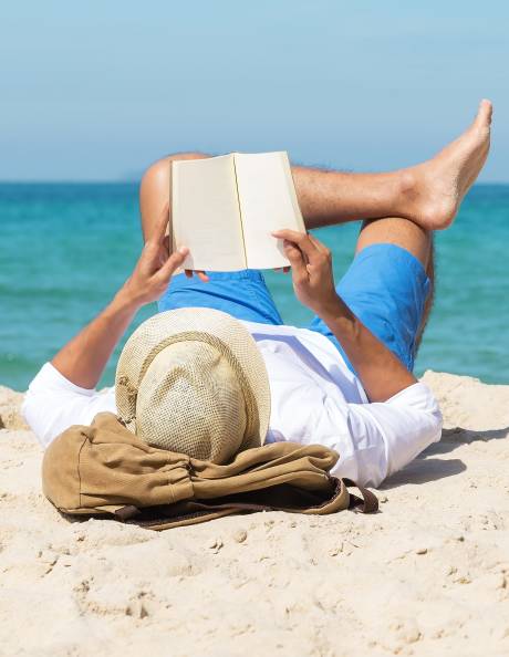 A man relaxes on a sandy beach, reading a book with the turquoise ocean in the background.