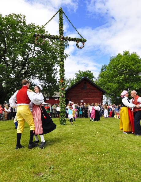 Dancers in traditional attire celebrate around a flower-adorned maypole in front of a rustic red barn.