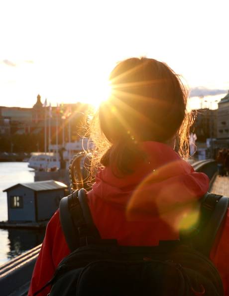 Golden sunlight streams through the hair of a traveler wearing a red jacket and backpack, overlooking a city skyline.
