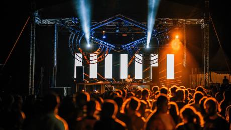 A DJ performs on a brightly lit stage in front of a large, silhouetted crowd at an outdoor music festival.
