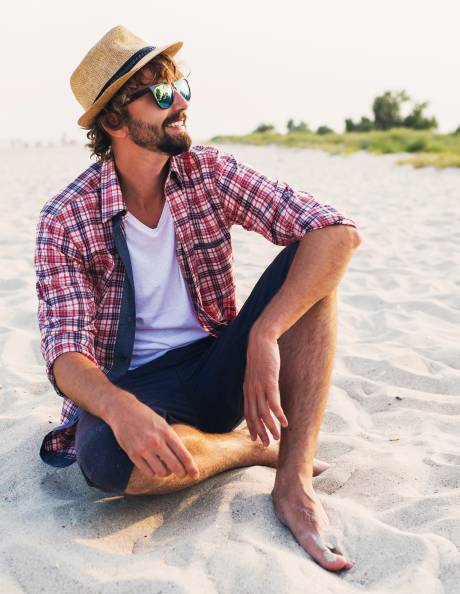 A man in a straw hat and plaid shirt relaxes on a sandy beach, enjoying the sun and sea breeze.