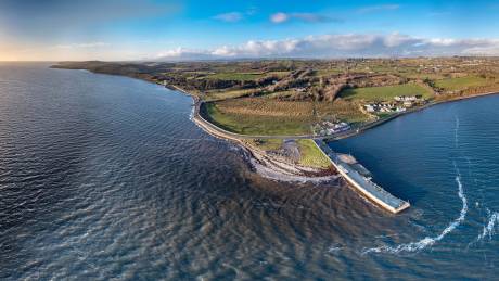 A concrete pier extends into the choppy blue waters of a bay, bordered by rolling green hills and a coastal road.