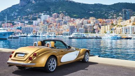 A gleaming gold convertible cruises along the waterfront in Monaco, with a stunning backdrop of yachts and colorful buildings.