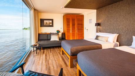 Two beds with dark grey blankets are neatly made in a room with a large window overlooking a wide river.