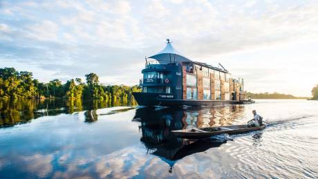 A man paddles a small canoe alongside the luxurious Aria Amazon cruise ship on a calm river.