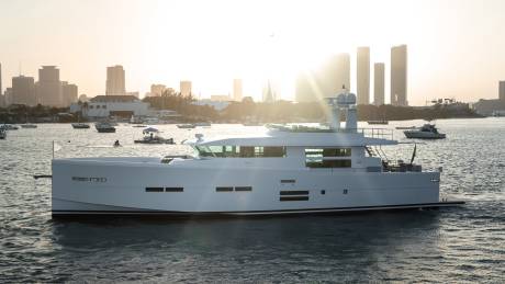 A sleek white yacht glides through the water with a city skyline bathed in golden sunlight in the background.