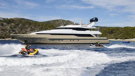 A luxury yacht anchors offshore as two jet skiers enjoy the crystal-clear waters of a Mediterranean island.