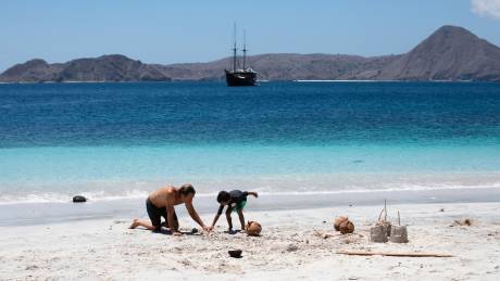 A father and son build sandcastles on a pristine beach with a traditional sailing ship anchored offshore.