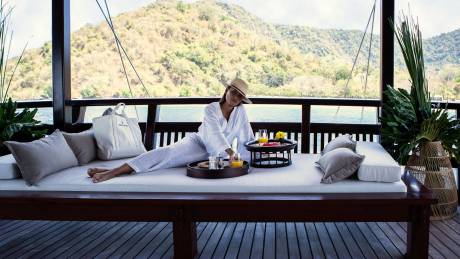 A woman in a white robe enjoys a leisurely breakfast on a daybed overlooking a lush, mountainous coastline.