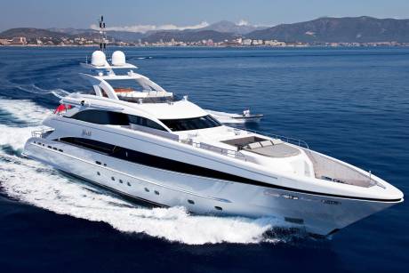 A sleek white yacht cuts through the deep blue sea, with a coastal town nestled against distant mountains in the background.