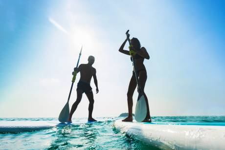 Silhouetted against a bright sun, a couple paddleboards across the gentle ocean waves.