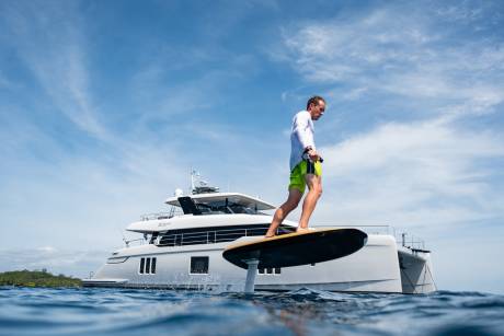A man in bright shorts rides an electric hydrofoil surfboard in front of a sleek catamaran yacht under a bright blue sky.