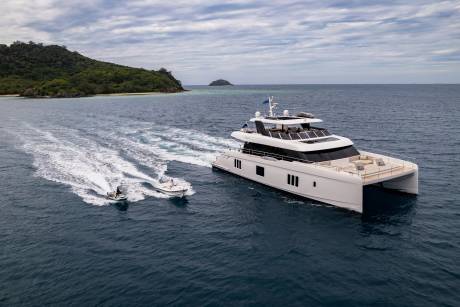 A luxurious catamaran yacht cruises through the ocean, trailed by a jet ski and a small tender near a lush, tropical island.