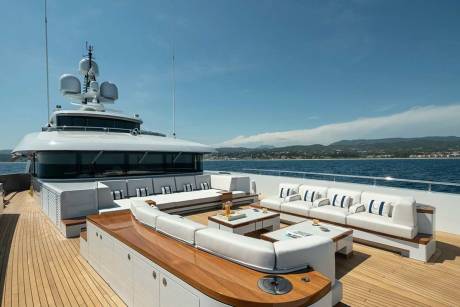 Luxurious white sofas and coffee tables create a stunning outdoor lounge on the deck of a yacht against a backdrop of the azure sea and distant hills.