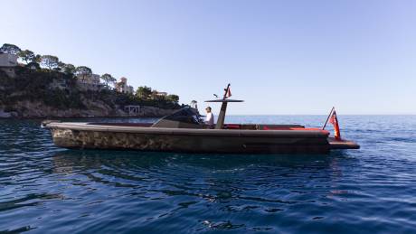 A man pilots a sleek, dark boat with red accents along a calm blue sea, with a cliffside village in the background.