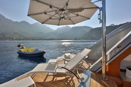 Relax on the deck of a luxury yacht under a large umbrella, with jet skis and a tender boat floating in the calm, sun-dappled water.