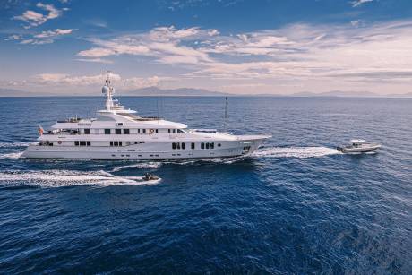 A luxurious white yacht cruises through the deep blue sea, trailed by a jet ski and a small tender boat.