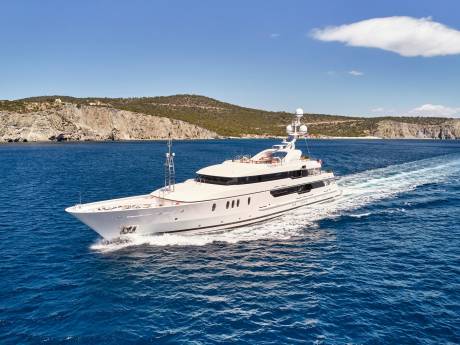 A sleek white yacht cruises through the azure waters off a rugged, verdant coastline under a clear blue sky.