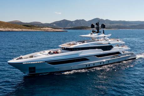 A sleek white yacht glides through the deep blue sea, with a rugged, tree-covered coastline in the background.