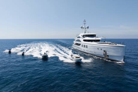 A sleek white superyacht cruises through the blue ocean, trailed by several jet skis and a small tender.