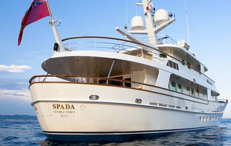 The luxurious yacht SPADA, flying a red ensign, cruises serenely on the calm blue sea under a clear sky.