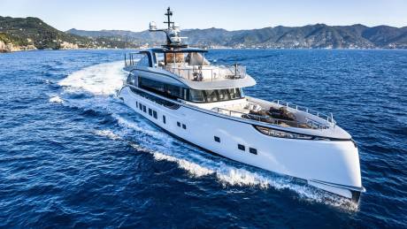A sleek white yacht cuts through the deep blue ocean with a mountainous coastline in the background.