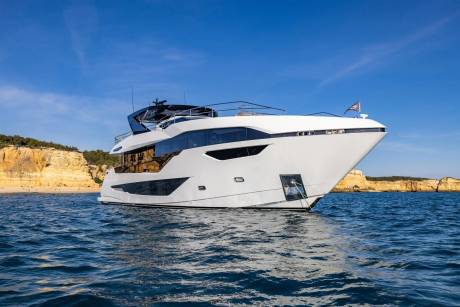 A sleek white yacht glides through the calm blue waters off a picturesque, sun-drenched coastline.