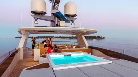 A couple enjoys drinks on a luxurious yacht deck with a sparkling turquoise hot tub and a serene coastal backdrop.