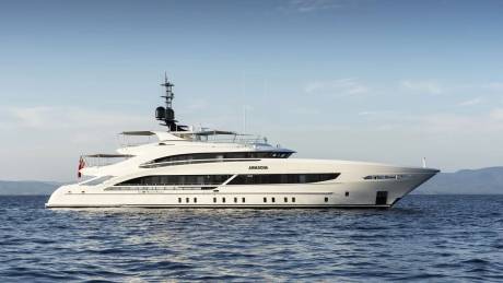 A large, luxurious white yacht is floating on the calm, blue waters of the ocean, with mountains visible in the background under a clear sky.